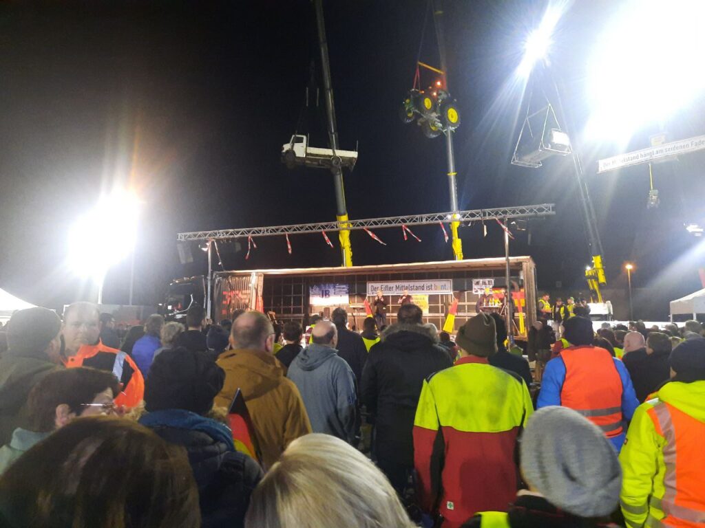 Bild aus der Demonstrantengruppe heraus auf die Bühne. 3 Krähne mit hochgezogenem Traktor, Lastwagen und PKW im Hintergrund.